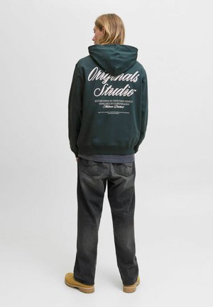 Eine Person mit blonden Haaren steht mit dem Rücken zur Kamera, trägt einen dunkelgrünen Kapuzenpullover mit weißem Text, dunkle Jeans und gelbe Stiefel vor einem schlichten Hintergrund.