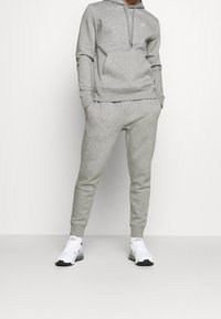 Felpa grigia in cotone con cappuccio e tasca frontale, abbinata a pantaloni jogger grigi con taglio affusolato. Scarpe da ginnastica bianche con dettagli neri.
