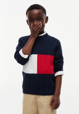 Dreng iført navyblå sweater med røde og hvide striber dækker sin mund med én hånd, stående foran en ensfarvet baggrund.