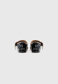 Un par de zapatos planos de charol negro con tirantes al tobillo y hebilla dorada, vistos desde atrás sobre un fondo blanco liso.