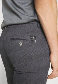 Domácí kalhoty s kostkovaným vzorem mají knoflíkovou zadní kapsu, malou nášivku s logem a texturovanou tkaninu. Pas obsahuje viditelný vícebarevný detail.