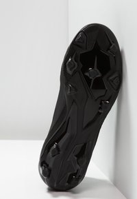 Black cleated soccer shoe sole angled against a white corner, showing textured studs and grip patterns for traction.