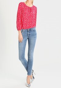 Blusa floral roja con mangas largas y escote recogido, combinada con jeans ajustados de color azul claro y zapatos de tacón alto plateados.