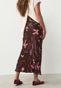 Jupe en satin marron ornée de motifs floraux rouges et roses ainsi que de rubans, présentant un design fluide et un ourlet légèrement évasé. Portée avec des ballerines bordeaux.