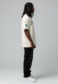 T-shirt à manches courtes de couleur crème avec des motifs graphiques verts, associé à un jean noir et des baskets blanches. Détails incluent une coupe décontractée.