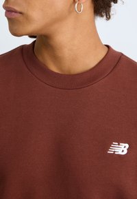 Brun bomullssweatshirt med ribbestrikket rund hals og liten hvit logo detajl. Stoffet ser glatt ut med en subtil tekstur.