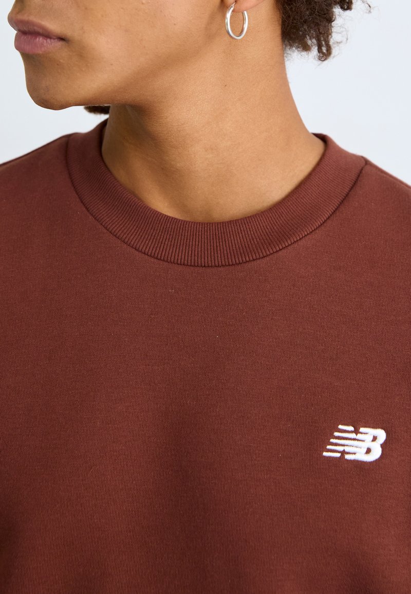 Brauner Baumwollpullover mit geripptem Rundhalsausschnitt und kleinem weißen Logodetail. Der Stoff wirkt glatt mit einer dezenten Textur.