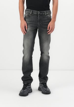 Personne portant un jean noir délavé et des chaussures de sport gris foncé, debout sur un fond blanc.