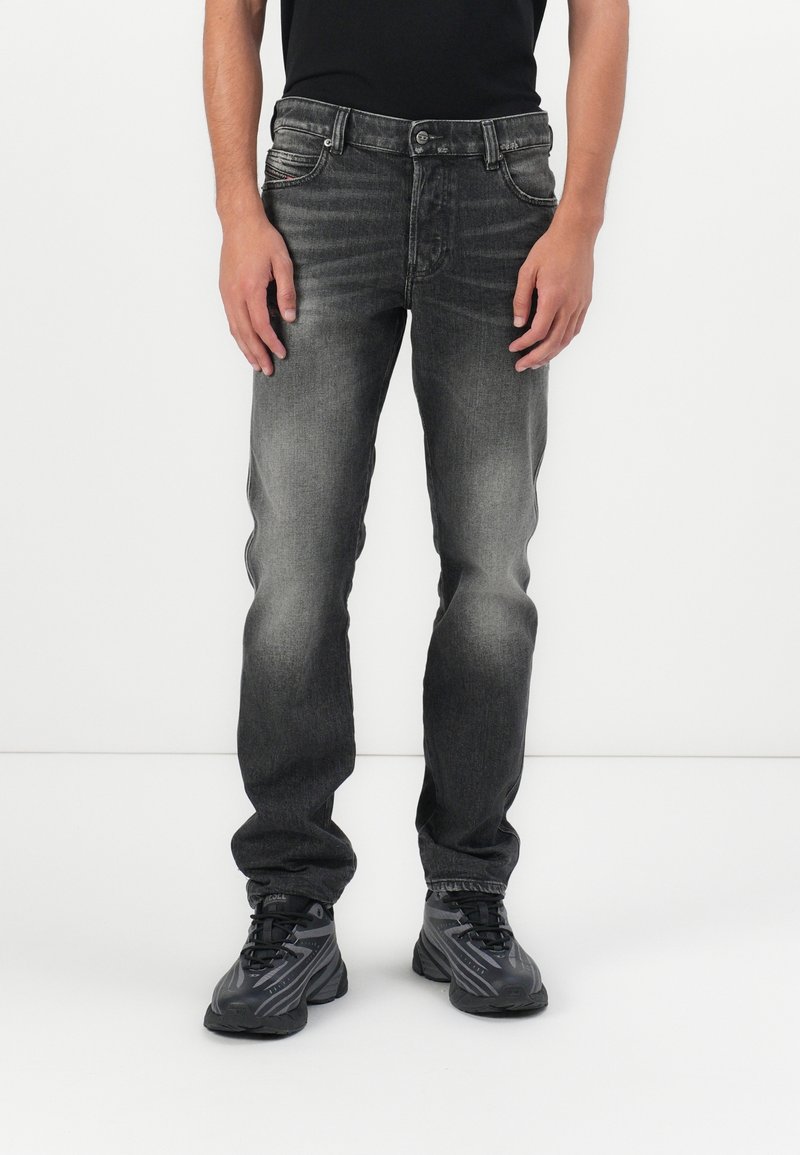 Personne portant un jean noir délavé et des chaussures de sport gris foncé, debout sur un fond blanc.