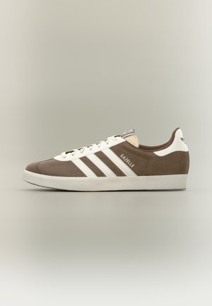 Καφέ και λευκό αθλητικό παπούτσι Adidas Gazelle με λευκές ρίγες, λευκά κορδόνια και υφασμένη ελαστική σόλα σε ουδέτερο φόντο.