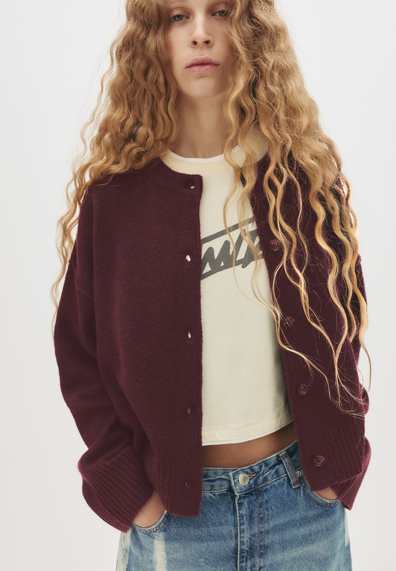 Cardigan bordeaux con polsini a coste e scollatura tonda, dotato di sei bottoni. Indossato sopra una camicetta corta color crema e jeans blu strappati.