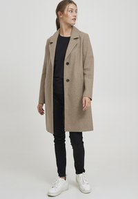 Cappotto di lana beige con colletto a revere, due bottoni neri e tasche laterali; abbinato a pantaloni neri e sneakers bianche.