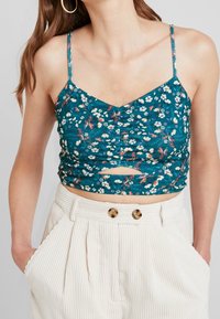 Blommig crop top i teal-färgad tyg med justerbara axelband, med en V-ringning och en front cut-out detalj. Texturerat mönster av små blommor.