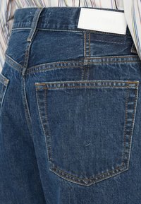 Τζιν μπλε υφάσματος με ψηλή μέση, πίσω τσέπες και αντίθεση πορτοκαλί ραφές. Ο ζωνάρι διαθέτει μια λευκή δερμάτινη ετικέτα με λογότυπο ανάγλυφο.