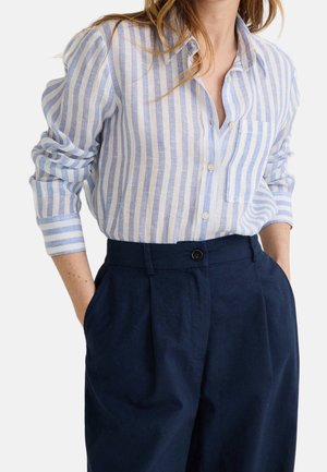 Kobieta nosząca koszulę z pionowymi jasnoniebieskimi i białymi paskami, włożoną w wysokie ciemnogranatowe spodnie, z rękami w kieszeniach.