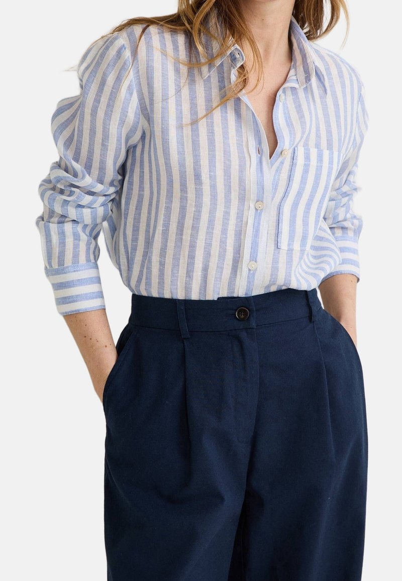 Femme portant une chemise à rayures verticales bleu clair et blanc, rentrée dans un pantalon taille haute bleu marine foncé, les mains dans les poches.