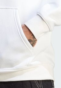 Felpa bianca con cappuccio e tasca a marsupio, maniche con polsini a costine. Visibile tatuaggio nero sul polso. Tessuto morbido e strutturato.