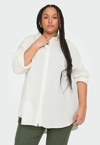 Chemise en lin blanc, ample, à manches longues et boutons, assortie à un pantalon vert. Texture douce et design décontracté.