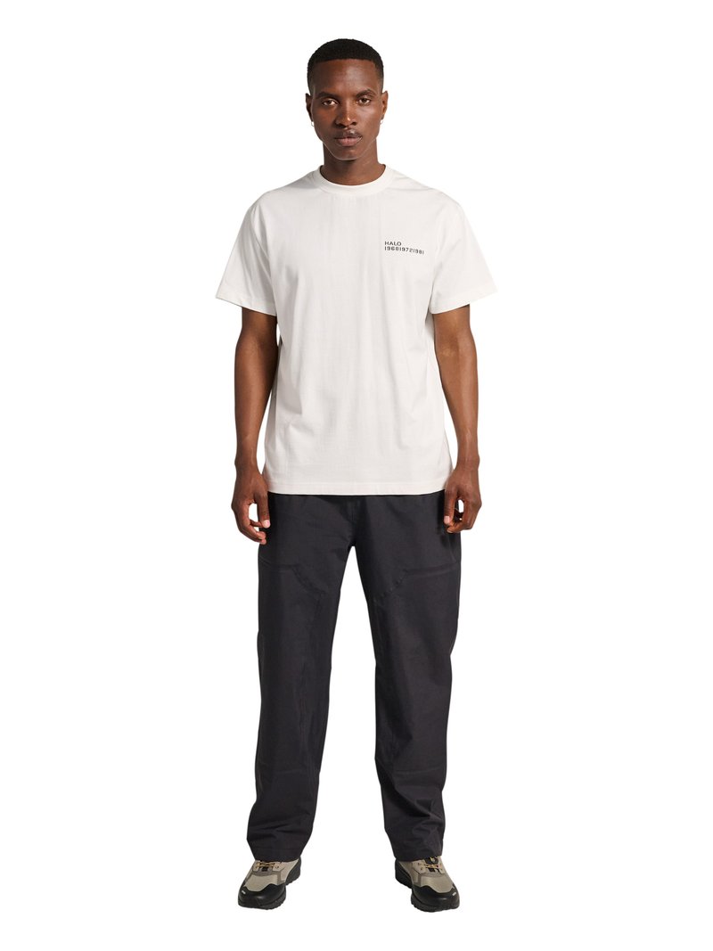 Mann steht mit entspannten Händen, trägt ein weißes kurzärmeliges T-Shirt mit Text auf der Brust, schwarze Hose und zweifarbige Freizeitschuhe.