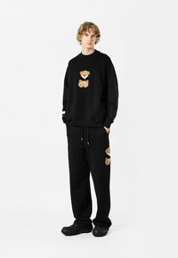 Conjunto de sudadera y pantalones negros con bordado de oso de peluche en tonos beige y blanco. La sudadera tiene un corte holgado; los pantalones tienen un cordón elástico.