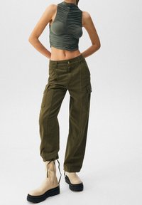 Pantaloni cargo verde olivo con tasche laterali, abbinati a un top senza maniche verde scuro con arricciature. Stivaletti beige alla caviglia con suola spessa.
