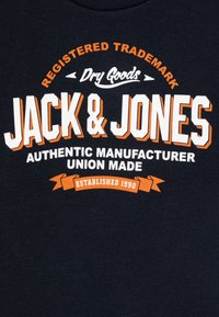 Jūras flotes kokvilnas t-krekls ar spilgti baltu un oranžu drukātu tekstu: "JACK & JONES," ar sīkāku informāciju "AUTENTISKS RAŽOTĀJS" un "IZVEIDOTS 1990".