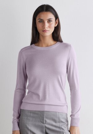 Kvinde med langt mørkt hår iført en lyse lilla crewneck-trøje og grå bukser, står op ad en ensfarvet hvid væg.