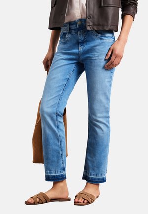 Femme portant un jean court bleu clair avec des ourlets effilochés, une veste en cuir marron, des sandales en daim beige et tenant un sac en daim marron.