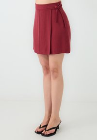 Bordeaux rok met plooien, zachte stof, strikdetail bij de taille en een rechte snit. G gedragen met zwarte slippers.