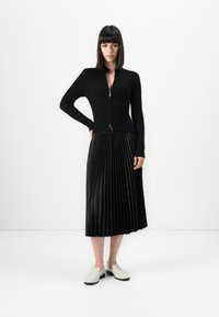 Woman standing with hand on hip wearing black zip-up sweater, black pleated midi skirt, and white slip-on shoes against white background.