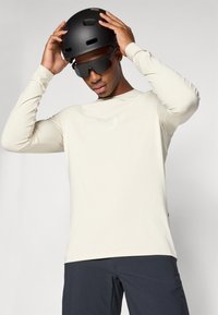 Pitkähihainen kerma V-neck -paita, jossa on huomaamaton logo, musta kypärä ja tummat aurinkolasit. Sileä kangas ja istuva muotoilu.