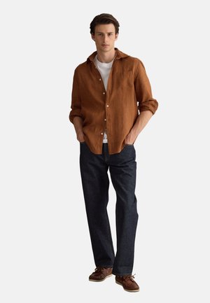 Jeune homme portant une chemise marron à boutons sur un t-shirt blanc, un jean foncé et des chaussures marron, debout avec les mains dans les poches.