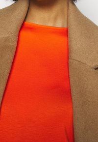 Brauner Wollmantel über einem leuchtend orangefarbenen, taillierten Oberteil. Der Mantel hat eine glatte Textur, während das Oberteil ein leicht geripptes Muster aufweist.