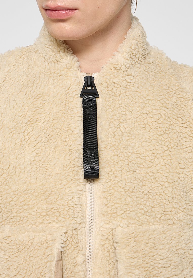 Persona che indossa una giacca in pile beige con un tirante nero della cerniera e due tasche frontali, mostrata dalla mento al petto.