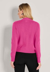 Eine Frau mit langen blonden Haaren trägt einen leuchtend pinkfarbenen Pullover mit langen Ärmeln und schwarze Hosen. Sie steht vor einer schlichten hellgrauen Wand.