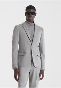 Traje gris claro con una chaqueta y pantalones a medida a juego. Presenta un corte ajustado, solapas de muesca y un bolsillo frontal. Llevado sobre un cuello de tortuga acanalado.
