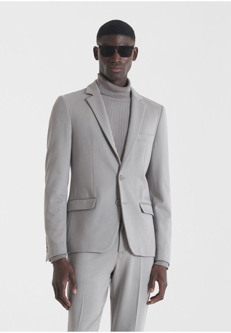 Traje gris claro con una chaqueta y pantalones a medida a juego. Presenta un corte ajustado, solapas de muesca y un bolsillo frontal. Llevado sobre un cuello de tortuga acanalado.