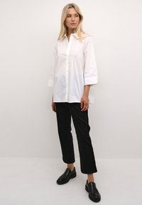Camicia bianca con bottoni, maniche lunghe, abbinata a pantaloni neri dritti e mocassini neri lucidi. Tessuto morbido e vestibilità rilassata.