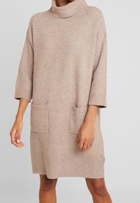 Abito in maglia di un morbido beige, con colletto alto, maniche a tre quarti e due tasche frontali. Vestibilità comoda, tessuto testurizzato.