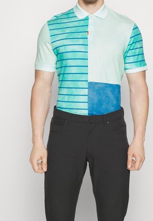 Man draagt een poloshirt met korte mouwen met een witte kraag, half effen en half blauw gestreept, gecombineerd met een zwarte broek.