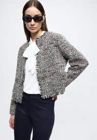 Jachetă din tweed cu fibre multicolore, cu un design scurtat și margini ciufulite, purtată peste o bluză albă și asortată cu pantaloni bleumarin.