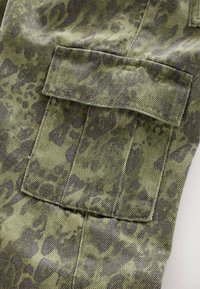 Krupan plan zelenog kamuflažnog materijala s velikim pravokutnim džepom sa šavom na površini.