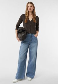 Dunkelbrauner, geknöpfter Cardigan mit langen Ärmeln, kombiniert mit lockeren, hellblauen weit geschnittenen Jeans. Das Model hält eine strukturierte, dunkle Handtasche.