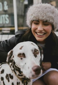 Dalmatinerhund med hvid pels og brune pletter, siddende ved siden af en person iført en fuzzy grå hat og sort jakke, som smiler.