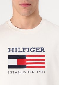 Bílé bavlněné tričko s vyšitým logem v námořnické modré, červené a bílé barvě s designem vlajky a textem "HILFIGER ESTABLISHED 1985".