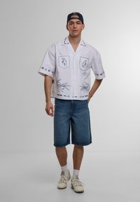 Witte shirt met korte mouwen en blauwe geborduurde details, gecombineerd met denim shorts. De outfit heeft twee borstzakken en opvallende bloemmotieven.