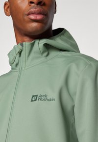 Green softshell jacket featuring a zip-up front, adjustable hood, and subtle logo. The fabric has a smooth texture with minimal seams.