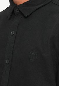 Svart skjorte med knapper og strukturert krage, laget av stoff med en subtil tekstur. Har svarte knapper og en liten brodert logo på brystet.