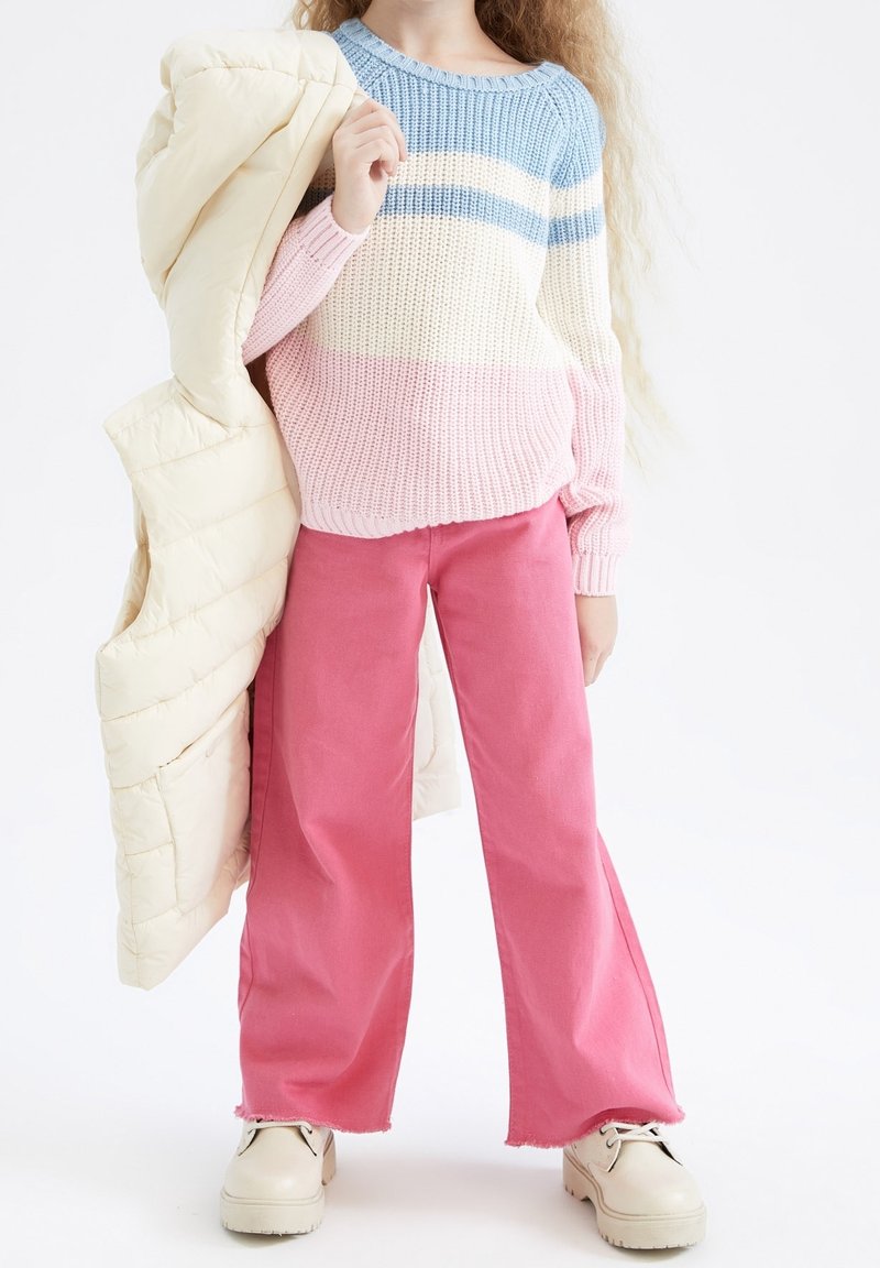 Enfant portant un pull à rayures pastel bleu, crème et rose, pantalon large rose, bottes crème, tenant une doudoune beige sur l'épaule.