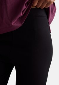 Černé legíny s hladkou texturou a přiléhavým střihem, zkombinované s volným bordeaux topem z prodyšného materiálu, částečně zastrčeným dovnitř.